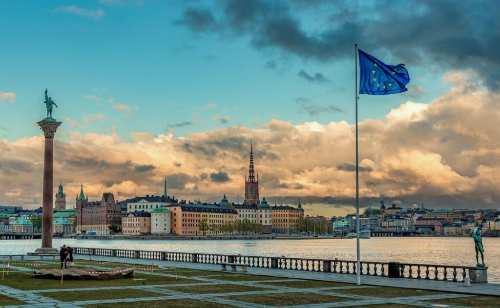 Stockholm waterfront view with iconic buildings and EU flag under a dramatic sunset sky.